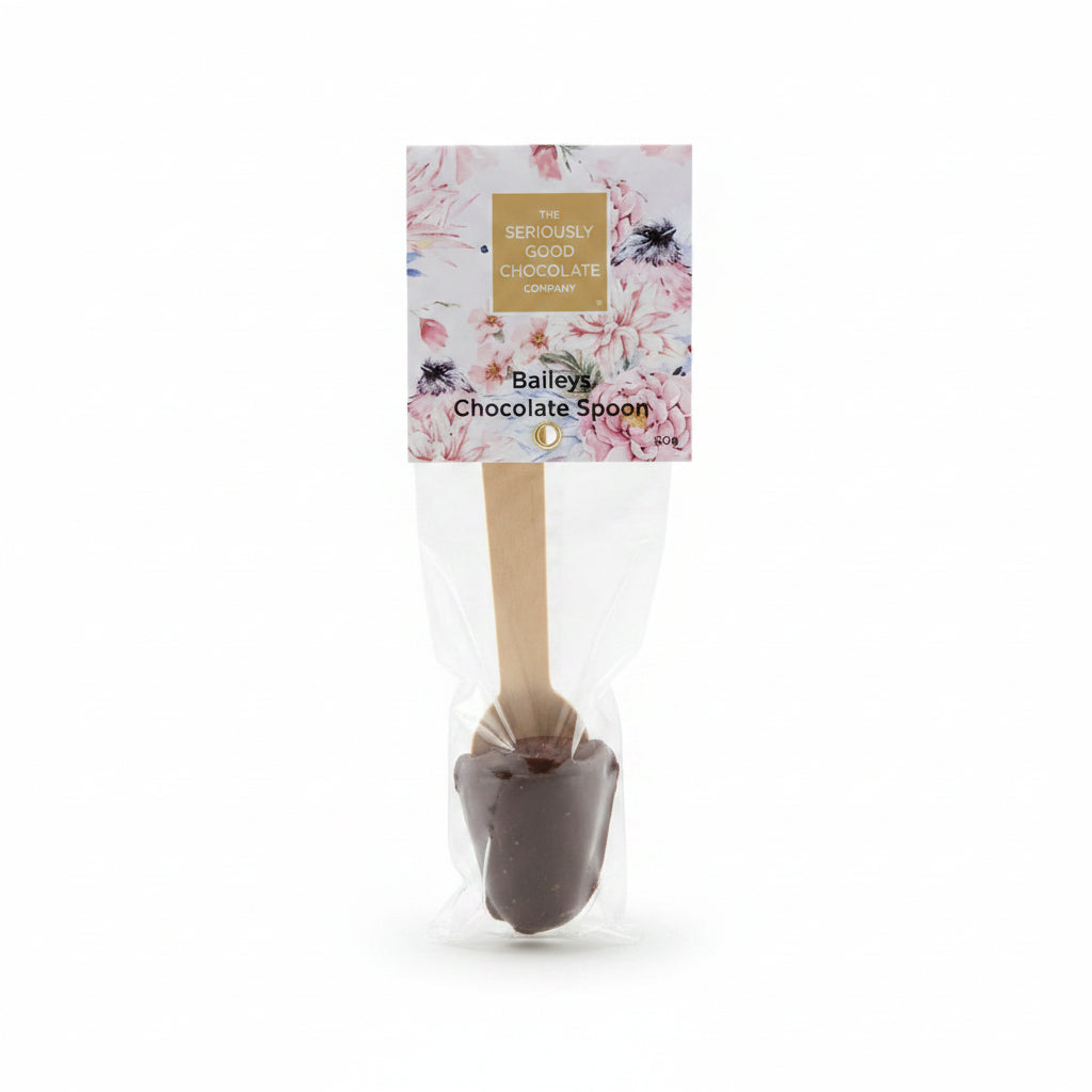 A packaged chocolate spoon with a floral label reading 'The Seriously Good Chocolate Company' and 'Bailey's Chocolate Spoon,' placed on a wooden surface surrounded by pink, red, and white flowers.
