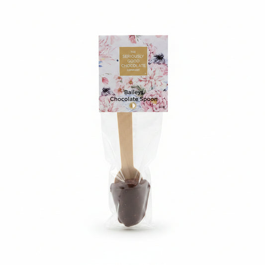 A packaged chocolate spoon with a floral label reading 'The Seriously Good Chocolate Company' and 'Bailey's Chocolate Spoon,' placed on a wooden surface surrounded by pink, red, and white flowers.