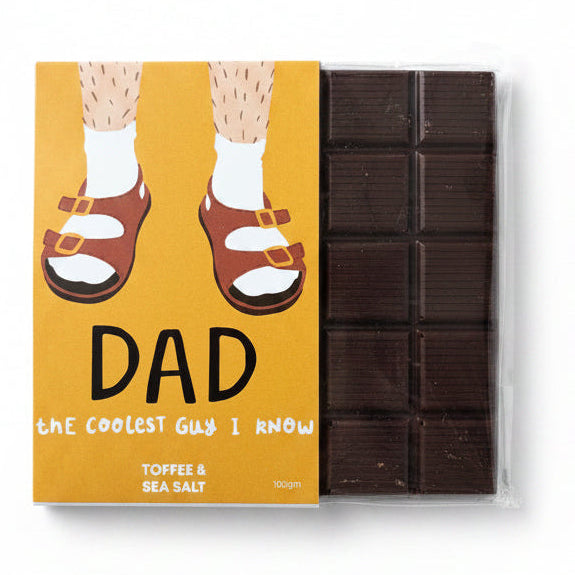 A packaged chocolate bar with a mustard-yellow printed label showing illustrated legs in brown sandals and white socks, next to the dark brown toffee and sea salt chocolate pieces in a clear plastic wrapper, on a light wood surface