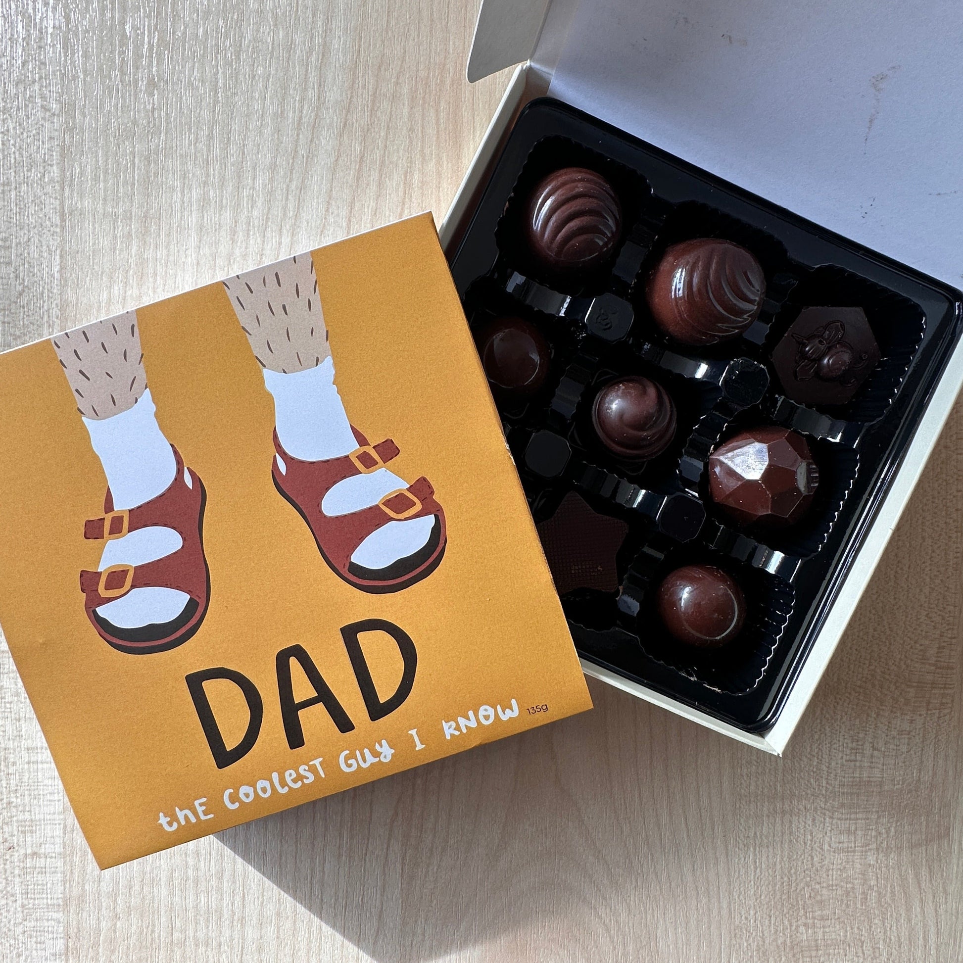 A box of chocolates with a humorous illustration of a dad's legs in a pair of red thongs on the cover, alongside a selection of individually wrapped chocolates with various toppings.