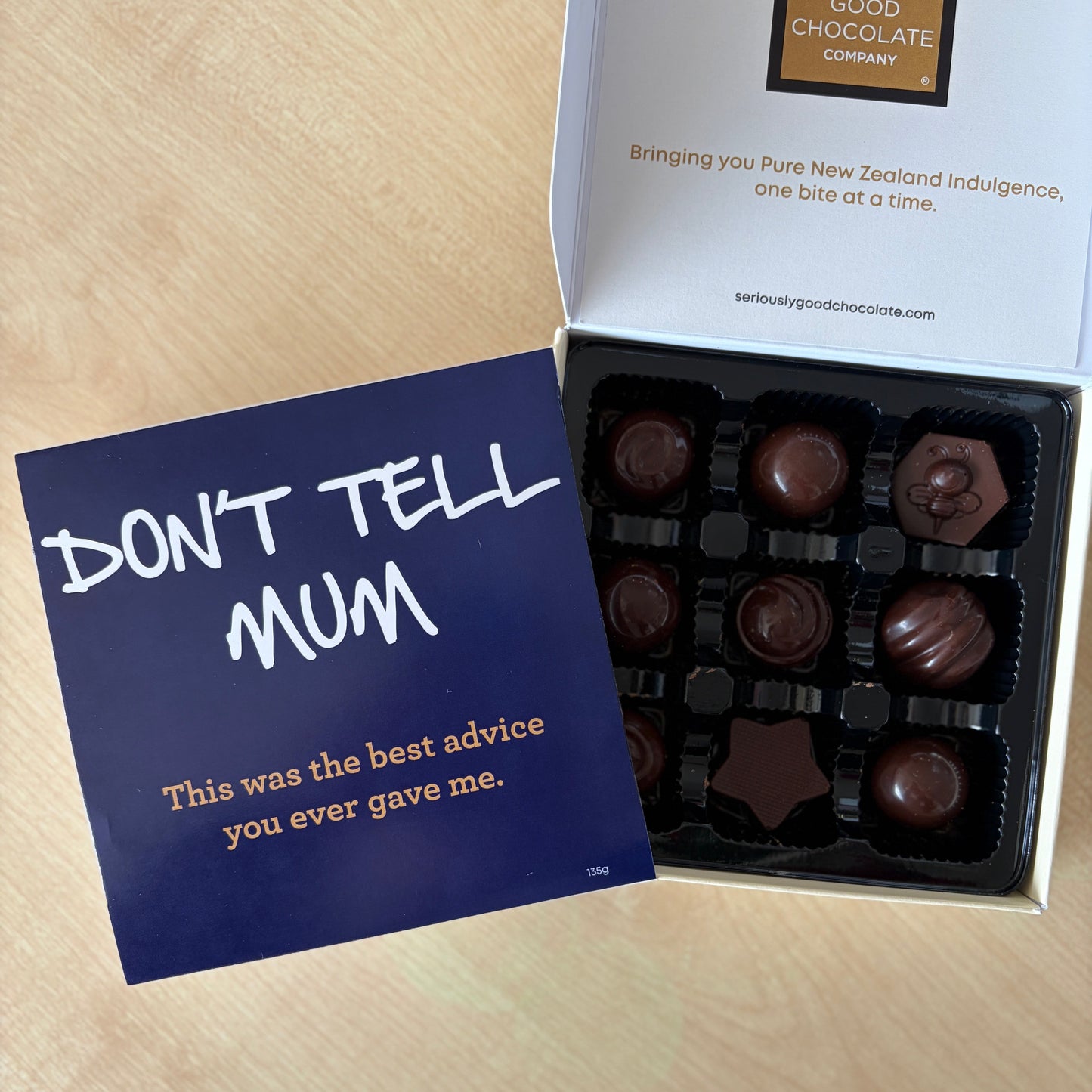 An open box of assorted chocolates on a light wood surface, with a dark blue card leaning against the box that reads “DON’T TELL MUM.”. Fathers day 2026