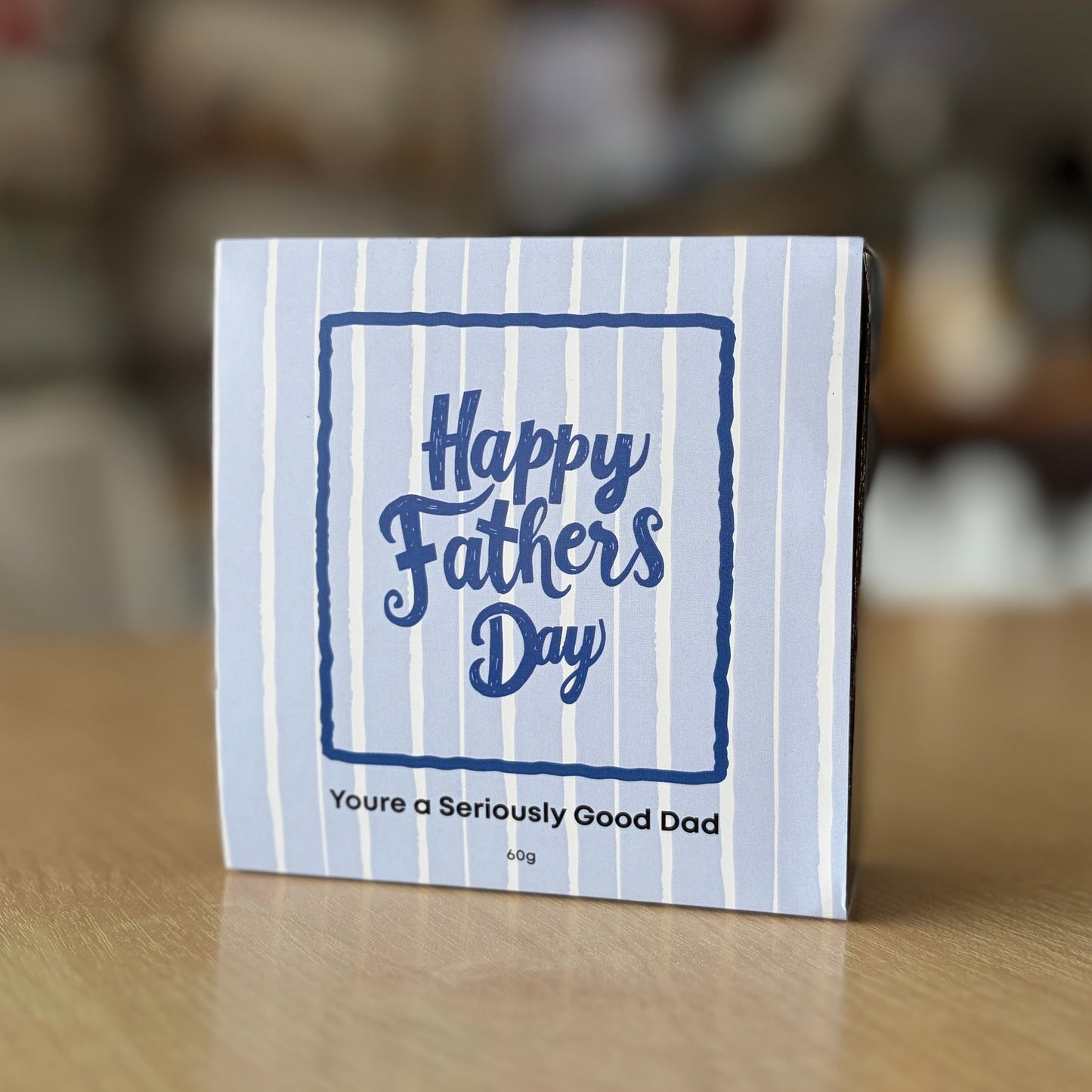 A small square Father's Day chocolate box with a blue and white striped design, standing on a wooden table in a blurred indoor setting. The front reads 'Happy Fathers Day' and 'You're a Seriously Good Dad'.