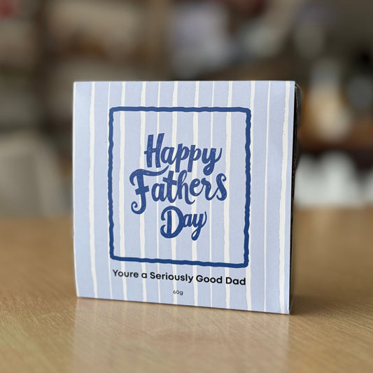 A small square Father's Day chocolate box with a blue and white striped design, standing on a wooden table in a blurred indoor setting. The front reads 'Happy Fathers Day' and 'You're a Seriously Good Dad'.