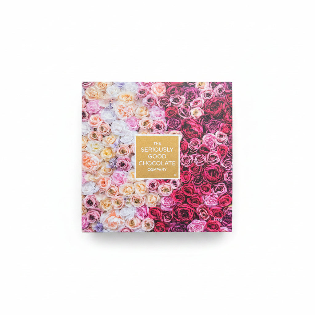 A square box of chocolates from The Seriously Good Chocolate Company, decorated with a dense pattern of multicolored roses in pink, red, yellow, and white, with a gold label in the center. The box is placed on a light wood surface decorated with pink and purple flowers in the background.