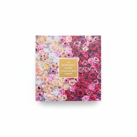 A square box of chocolates from The Seriously Good Chocolate Company, decorated with a dense pattern of multicolored roses in pink, red, yellow, and white, with a gold label in the center. The box is placed on a light wood surface decorated with pink and purple flowers in the background.