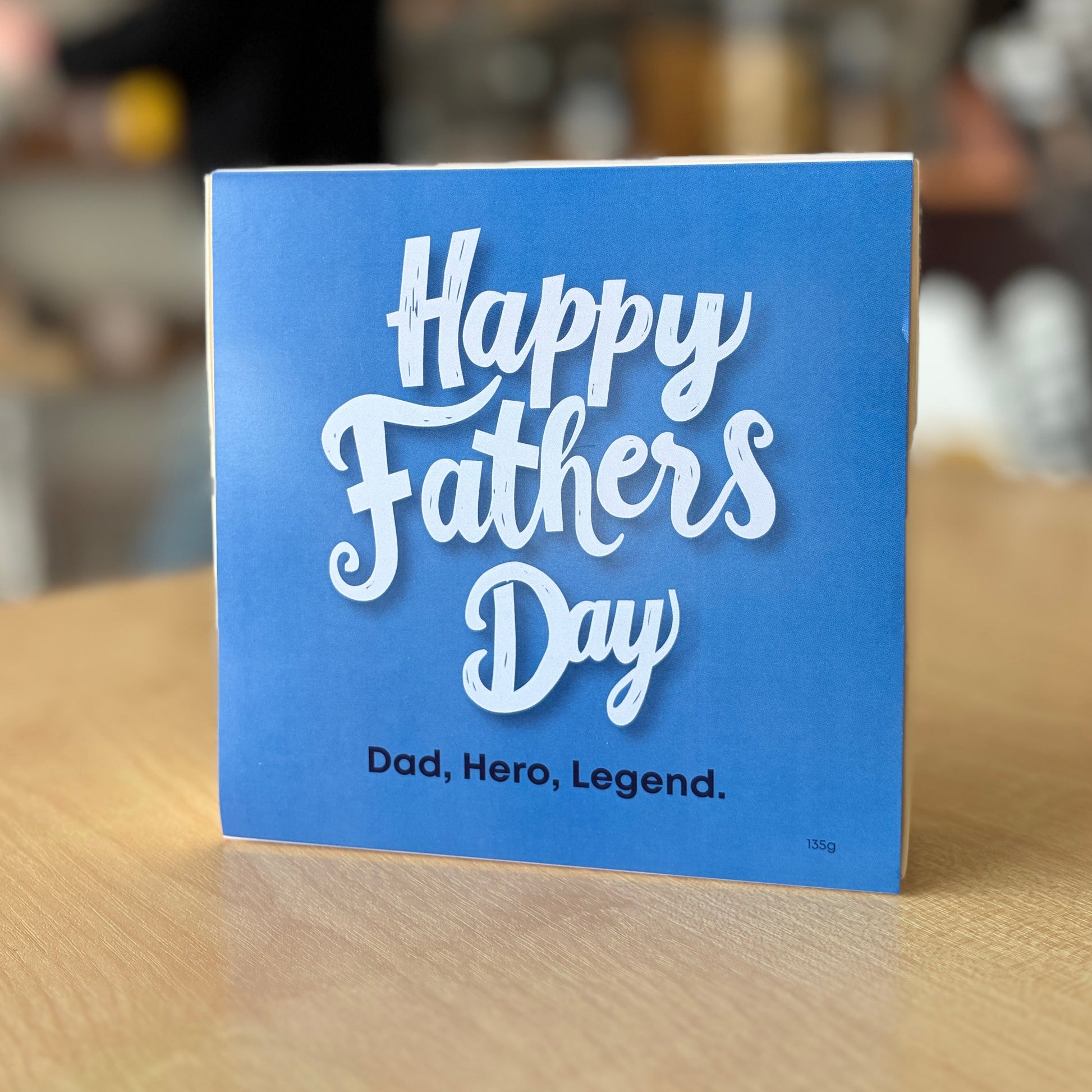 A square blue chocolate box with white text reading 'Happy Fathers Day' and 'Dad, Hero, Legend.' standing on a light wood table in a blurred indoor setting.