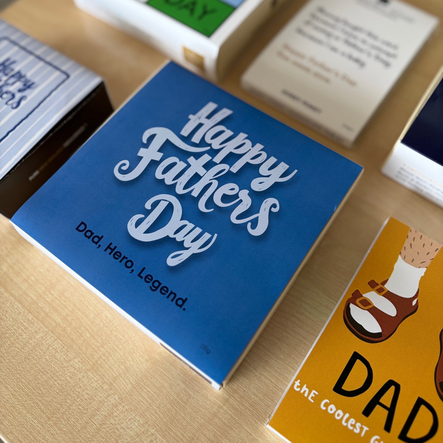 A blue square chocolate box with large white script reading 'Happy Fathers Day' and smaller text 'Dad, Hero, Legend.' sits on a light wood surface, surrounded by other Father's Day themed chocolate boxes
