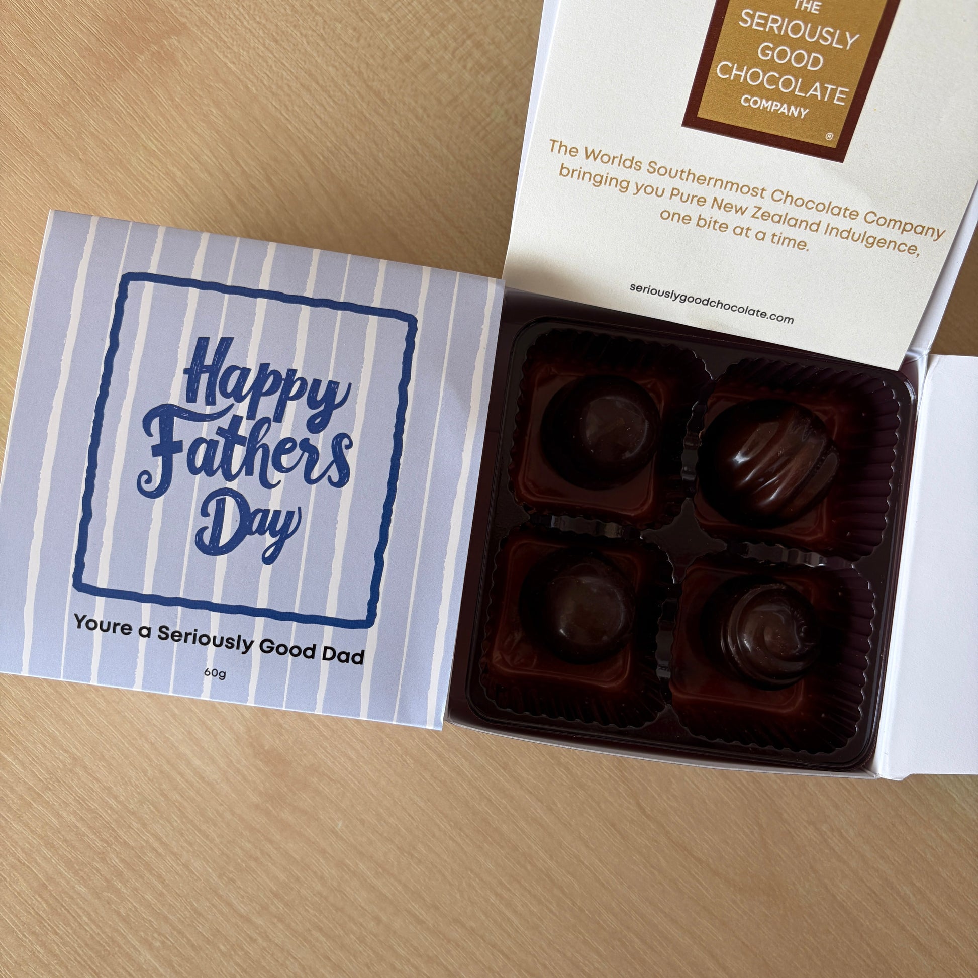 An open gift box of chocolates on a light wood surface, with the lid printed 'Happy Saffron Day' and the box labeled 'The Saffronly Chocolate Company.' Four dark chocolate pieces are visible inside a white box.