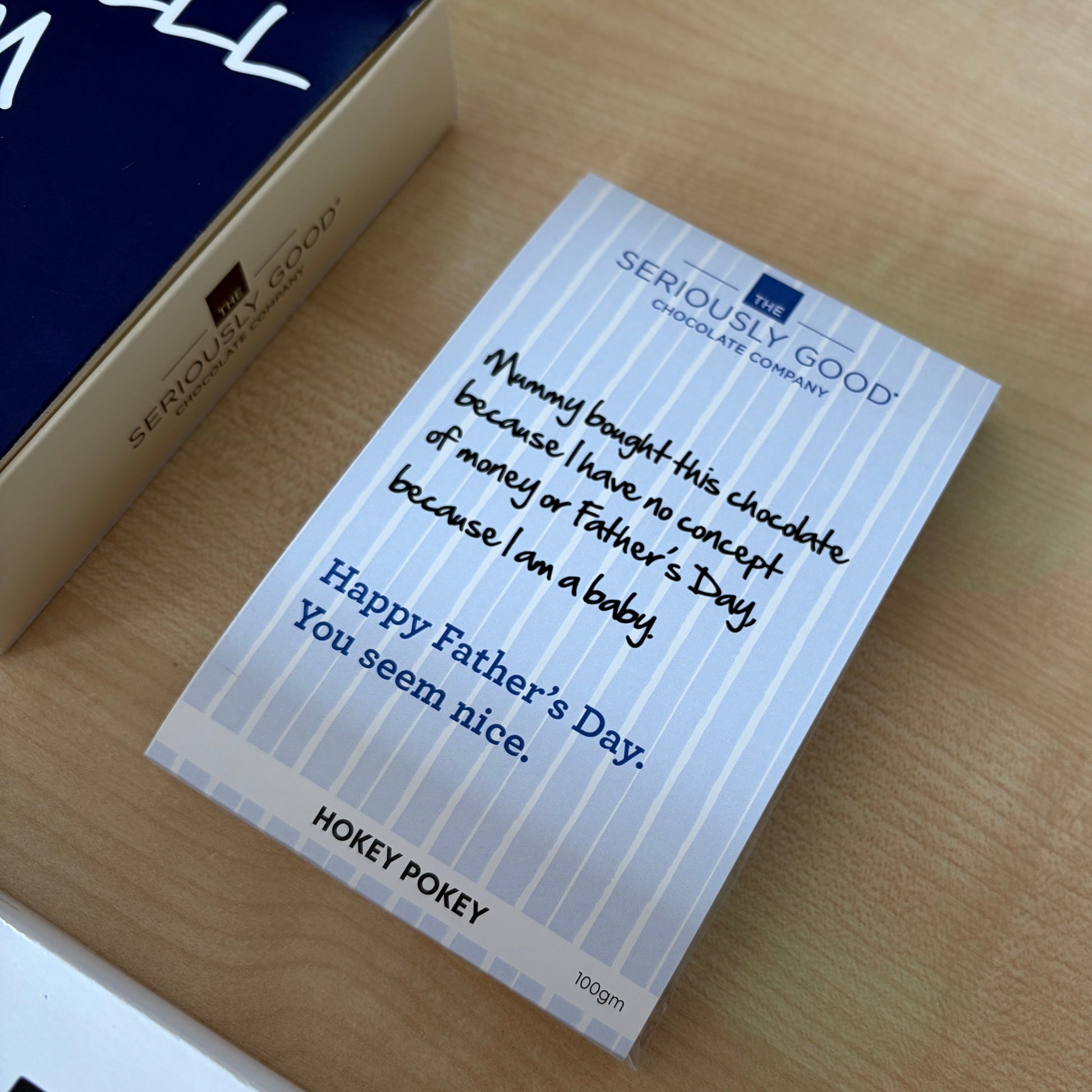 A small rectangular chocolate block from Seriously Good, featuring a light blue and white striped design with a handwritten message for Father's Day, placed on a light wood tabletop next to its dark blue and gold box.