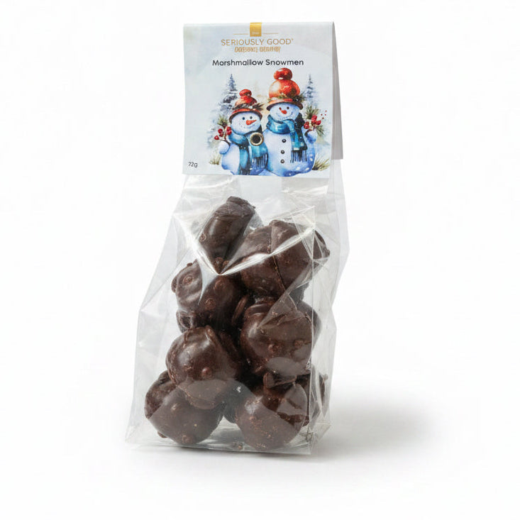 A bag of Seriousy Good's Marshmallow Snowmen chocolate treats sits on a wooden table next to a decorative nutcracker and holiday ornaments. The packaging features a white card with illustrations of two snowmen.