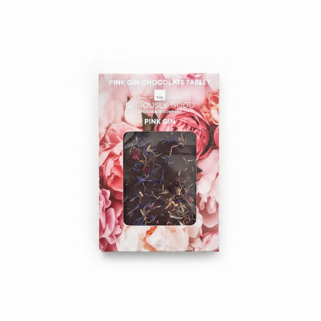 A boxed pink gin chocolate tablet from Seriously Good Chocolate Company, featuring a floral pink rose design and a clear window showing dark tablets inside, photographed on a light wood surface with flowers around it.