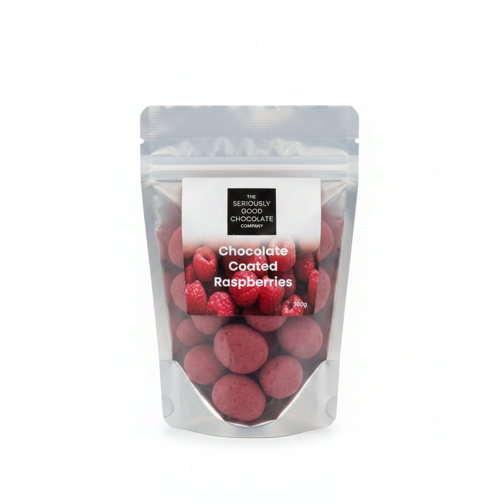 A clear stand-up pouch containing red chocolate-coated raspberries, standing on a wooden table with a blurred cafe interior in the background.