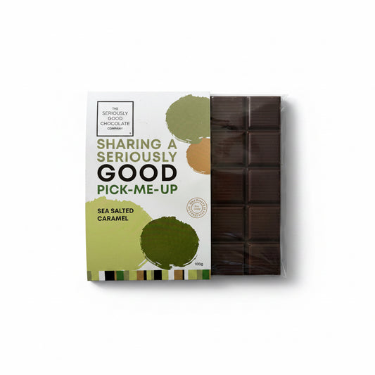 A packaged bar of chocolate from The Seriously Good Chocolates on a light wood tabletop. The white and green wrapper reads 'Sharing a Seriously Good Pick-Me-Up' and 'Sea Salted Caramel.