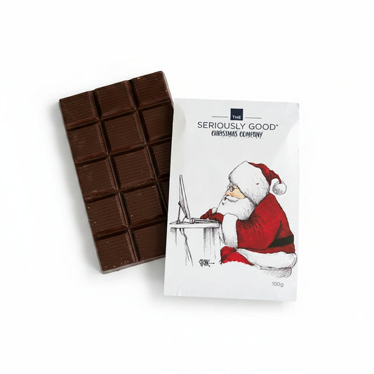 A packaged chocolate block with 'Seriously Good Christmas Company' branding, depicting Santa Claus on the front, placed on a wooden surface surrounded by Christmas decorations including a faux fur hat and golden confetti.