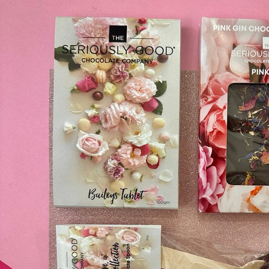A flat-lay of The Seriously Good Chocolate Company's Pink Gin Chocolate Collection, featuring a large floral-patterned chocolate tablet box, a smaller pink floral booklet, and a chocolate dip with a wooden spoon in clear wrapping, arranged on a pink and rose gold background.