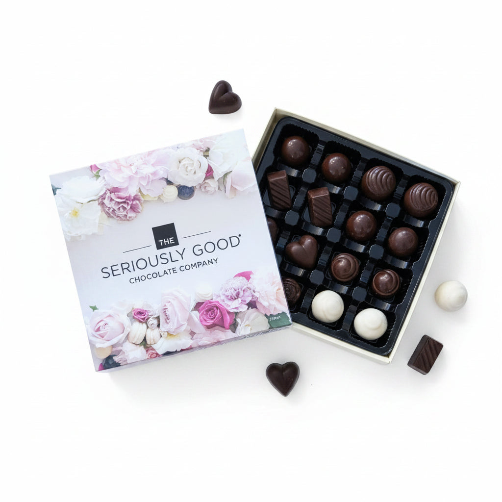 An open box of chocolates from The Seriously Good Chocolate Company sits on a wooden table, surrounded by scattered flower petals, flowers, and a few loose chocolates. The box lid shows a floral design with the brand name, and the open tray contains assorted milk and dark chocolate pieces.