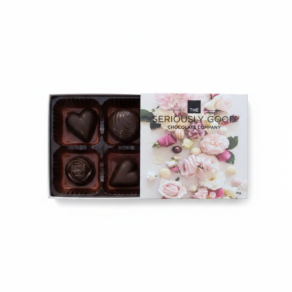 An open box of chocolates from The Seriously Good Chocolate Company on a wooden surface, surrounded by pink flower petals and dark chocolate pieces. The box lid features a floral design with the brand name.