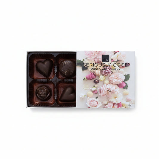An open box of chocolates from The Seriously Good Chocolate Company on a wooden surface, surrounded by pink flower petals and dark chocolate pieces. The box lid features a floral design with the brand name.