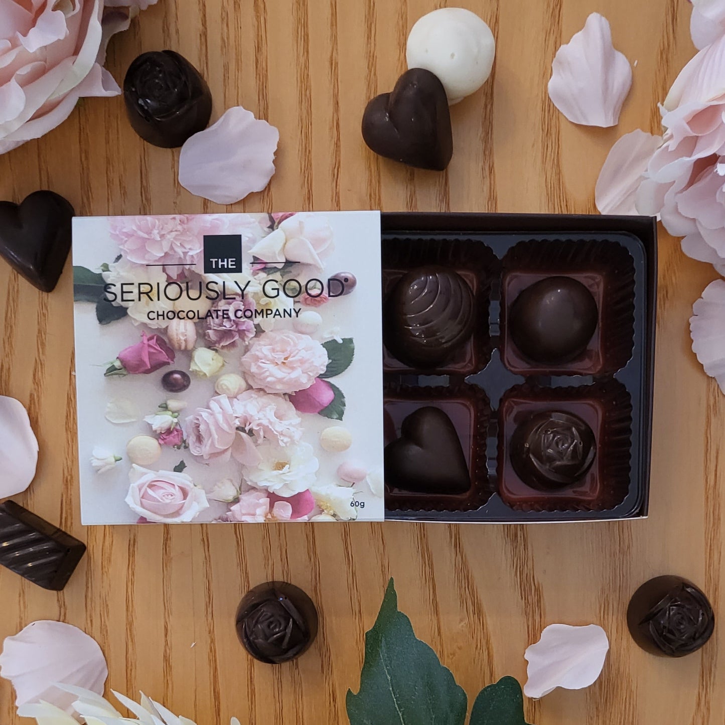 A box of chocolates from The Seriously Good Chocolate Company, presented with an assortment of chocolate shapes including hearts and domes, displayed on a wooden surface with floral accents.