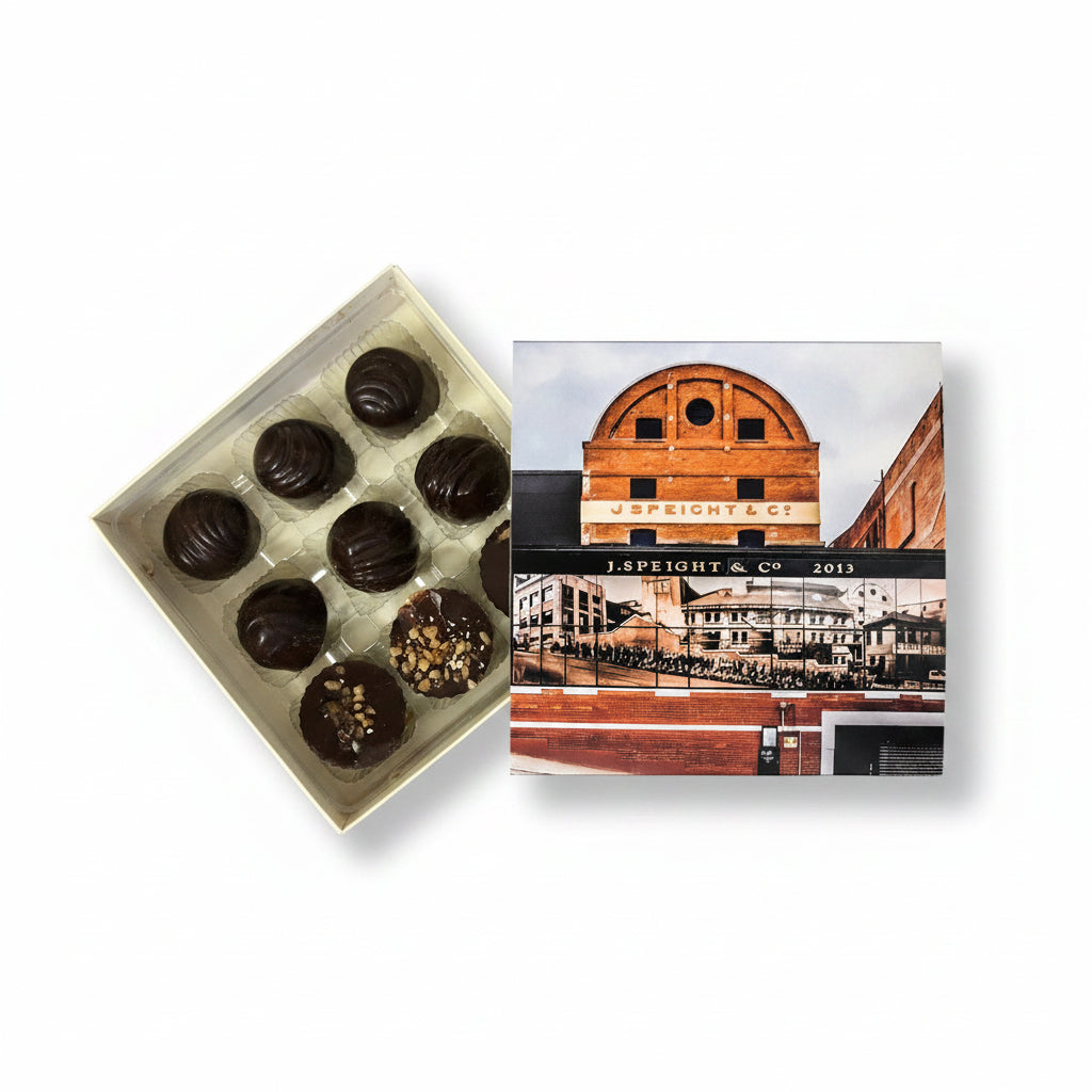 A box of Speight's beer infused chocolates with nine pieces. The chocolates are dark and appear to be covered in a nut topping, placed in front of a cardboard box with a printed image of a building.