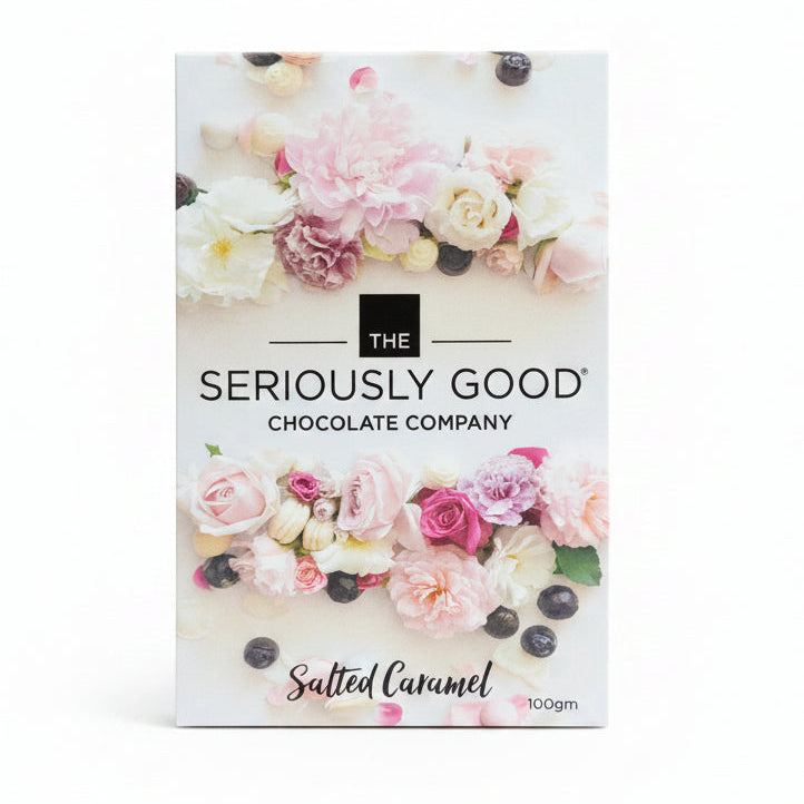 A chocolate block wrapped in clear packaging, with a floral design packaging box branded 'The Seriously Good Chocolate Company, Salted Caramel'. Presented on a wooden surface with pink flowers in the background.