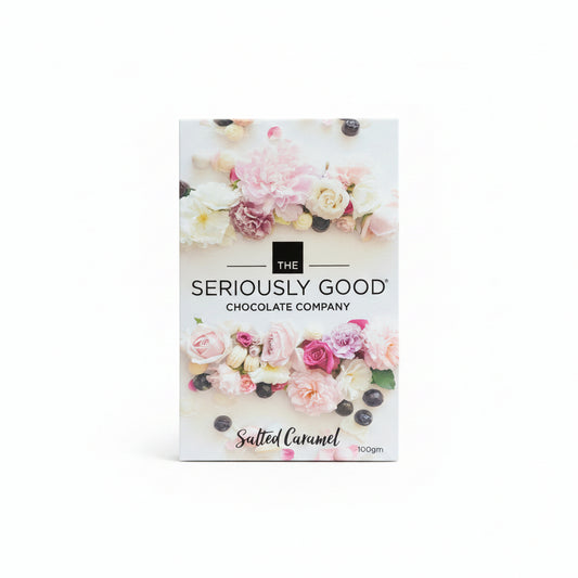 A chocolate block wrapped in clear packaging, with a floral design packaging box branded 'The Seriously Good Chocolate Company, Salted Caramel'. Presented on a wooden surface with pink flowers in the background.
